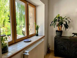 Ein Zimmer mit Fensterblick auf einen Garten, einem Heizkörper, einer Blumentopfpflanze und einem Schrank mit dekorativen Schnitzereien.