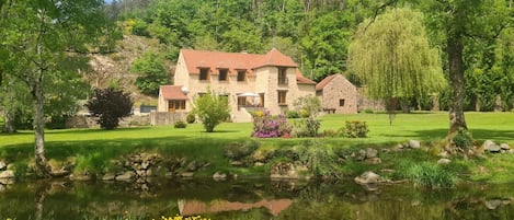 A stone house with a red roof, surrounded by a pond, trees, and a grassy lawn.