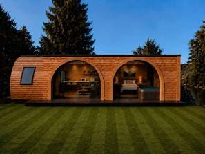 A modern, compact house with a brick exterior, two arched entryways, a visible kitchen area, and a bedroom with a bed and seating. - The Bridge Collection (Brandon)