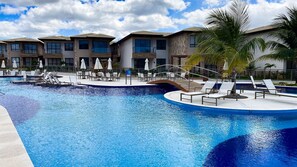 A swimming pool with lounge chairs and umbrellas, surrounded by multi-story buildings with balconies. - Casa em Resort c 4 Su tes na Praia de Guarajuba (Camaçari)