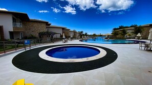A swimming pool area with a circular design, surrounded by lounge chairs and umbrellas, and a stone building with a balcony. - Casa em Resort c 4 Su tes na Praia de Guarajuba (Camaçari)