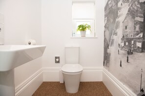 A bathroom with a toilet, a sink, a window with a plant, and a wall-mounted toilet paper dispenser. - Eastfield - Half House (Leyburn)