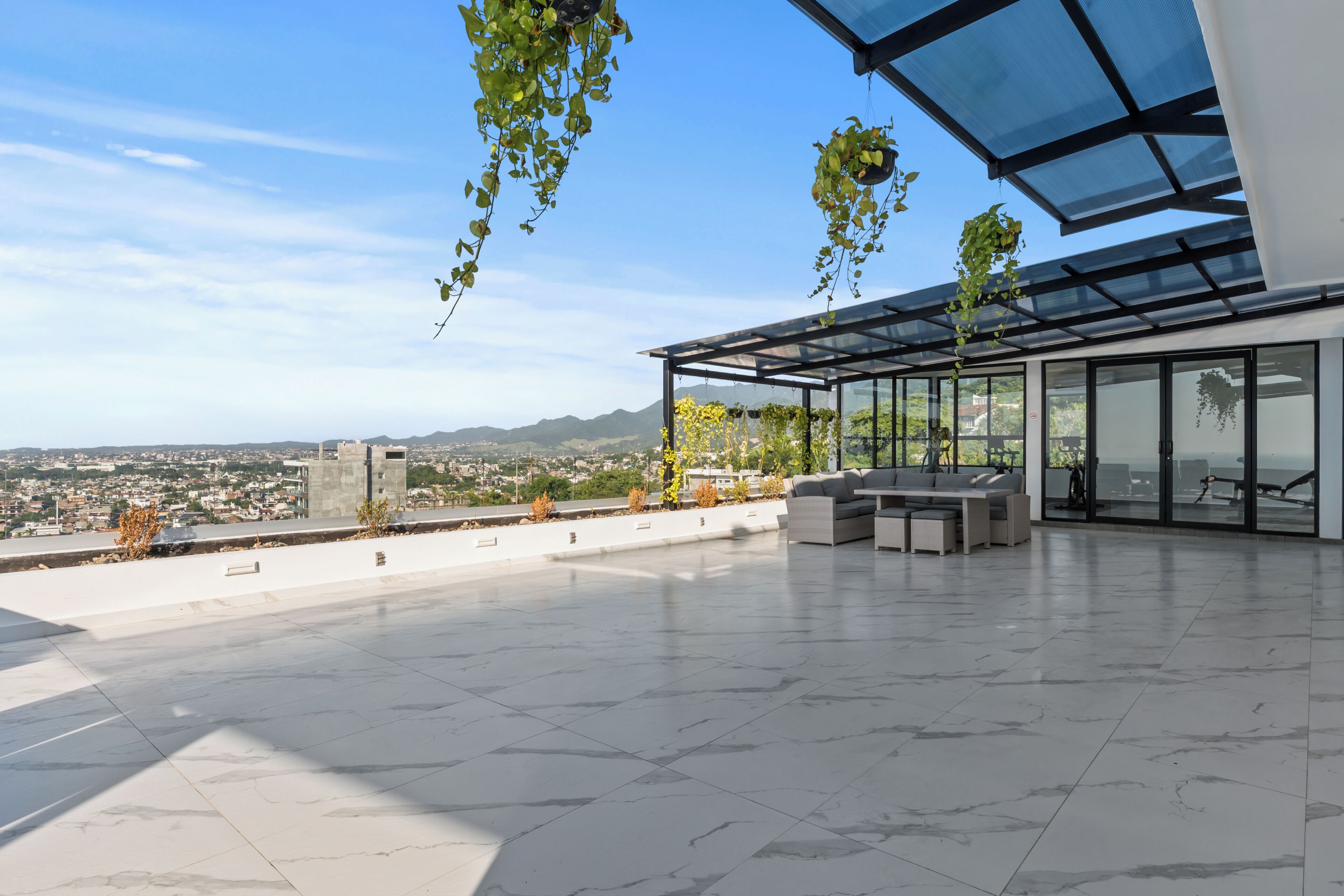 A rooftop terrace with a glass enclosure, outdoor furniture, and hanging plants.