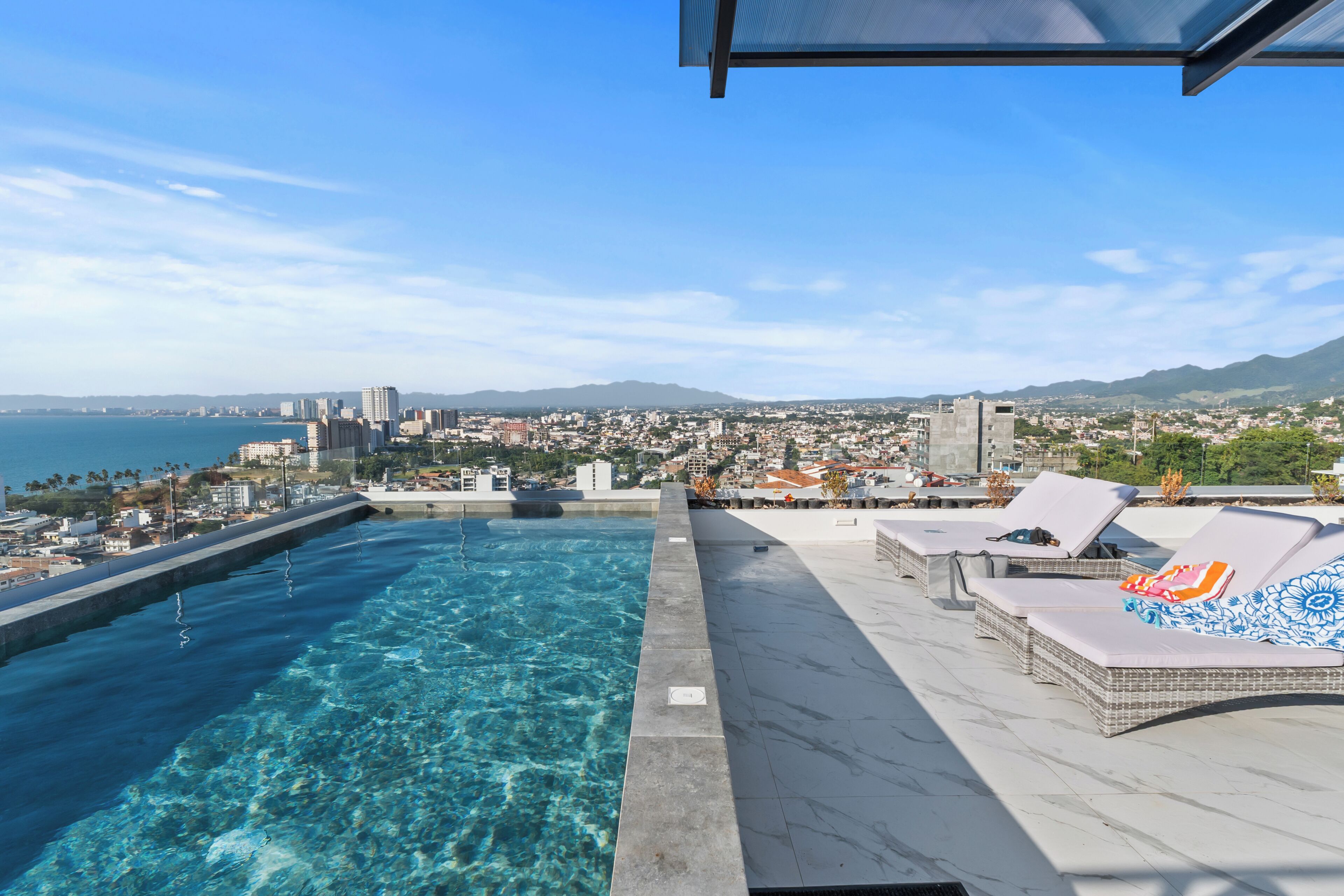 A rooftop pool with clear blue water, lounge chairs, and a view of a cityscape and mountains in the distance.