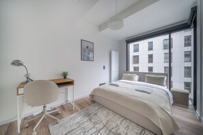 Ein Schlafzimmer mit einem Bett, einem Schreibtisch, einem Stuhl, einem Fenster und Blick auf ein Gebäude.