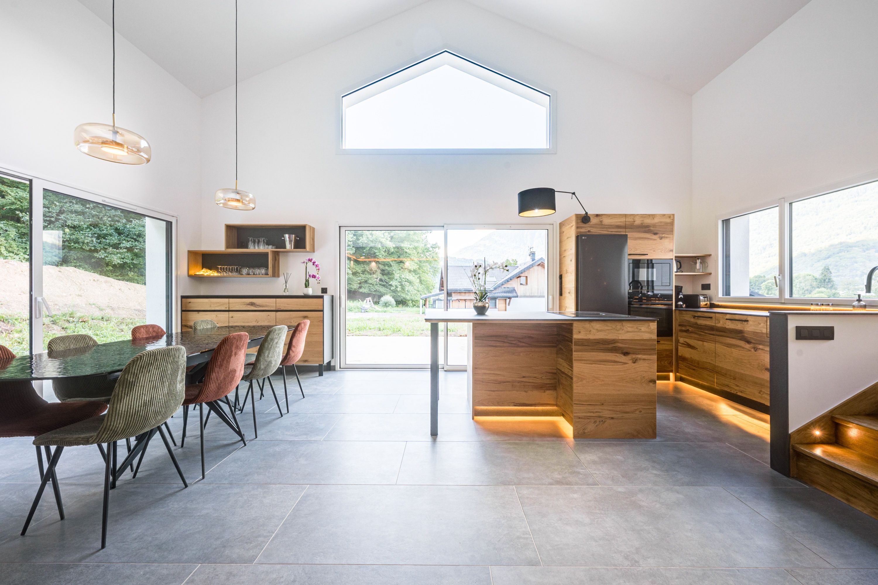 A modern kitchen with a central island, wooden cabinetry, and a large window overlooking a garden.
