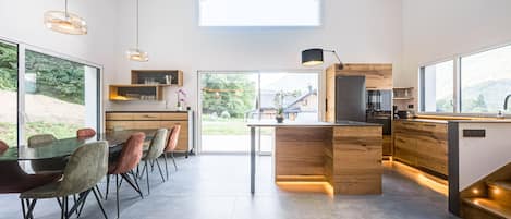 A modern kitchen with a central island, wooden cabinetry, and a large window overlooking a garden.