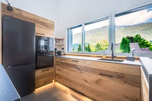 Eine moderne Küche mit Holzschränken, einem schwarzen Kühlschrank und einem großen Fenster mit Blick auf eine gebirgige Landschaft.
