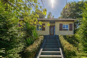 Uma casa com entrada frontal, escadas que levam à porta e um jardim com diversas plantas.