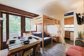 A compact room with a bunk bed, a dining table with chairs, a small sofa, a kitchen area, and large windows. - La Pause du Lindar - Studio avec Balcon (Aillon-le-Jeune)