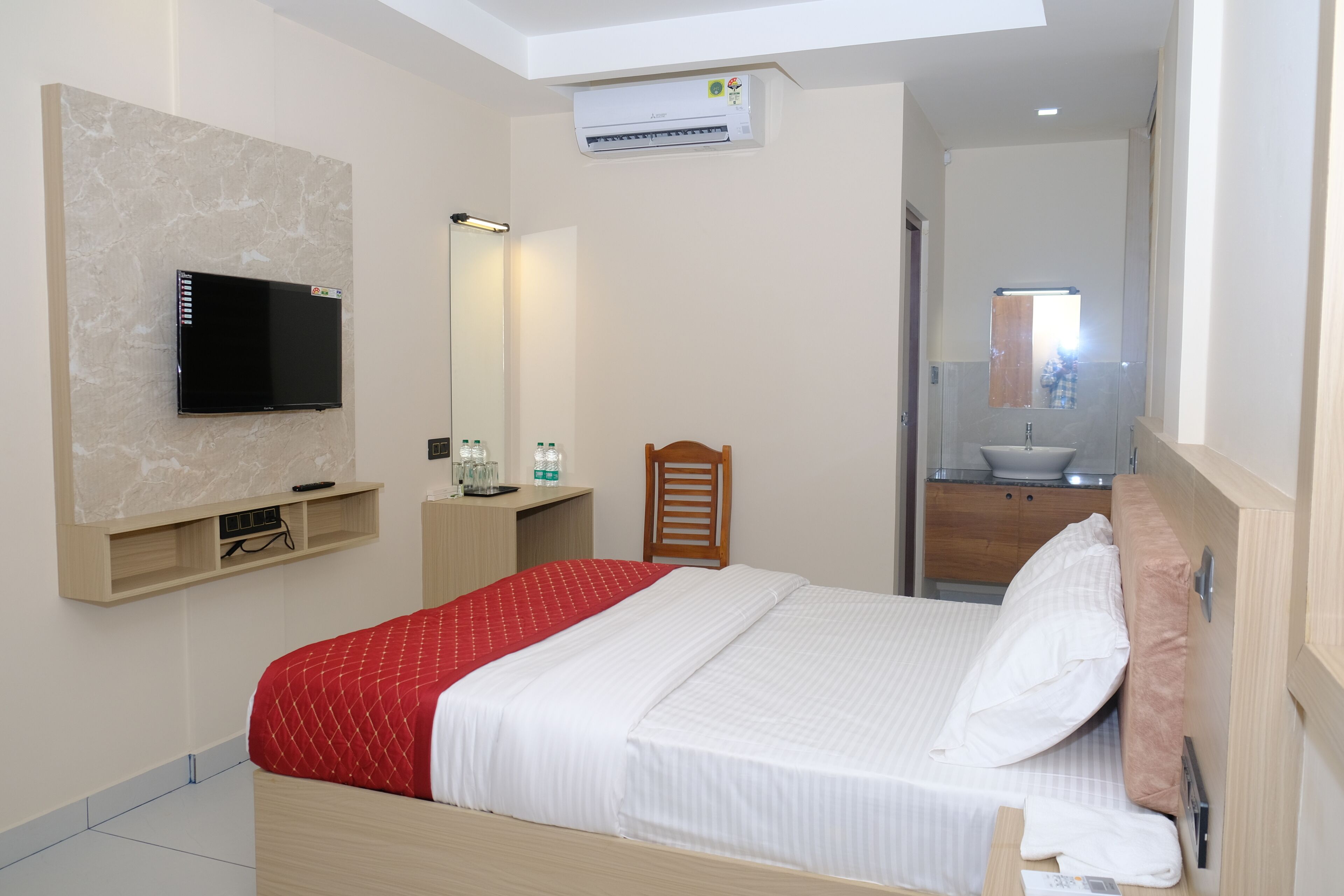 A hotel room with a bed, a television mounted on the wall, a bedside table with a remote control, a mirror, and a bathroom with a sink and mirror.