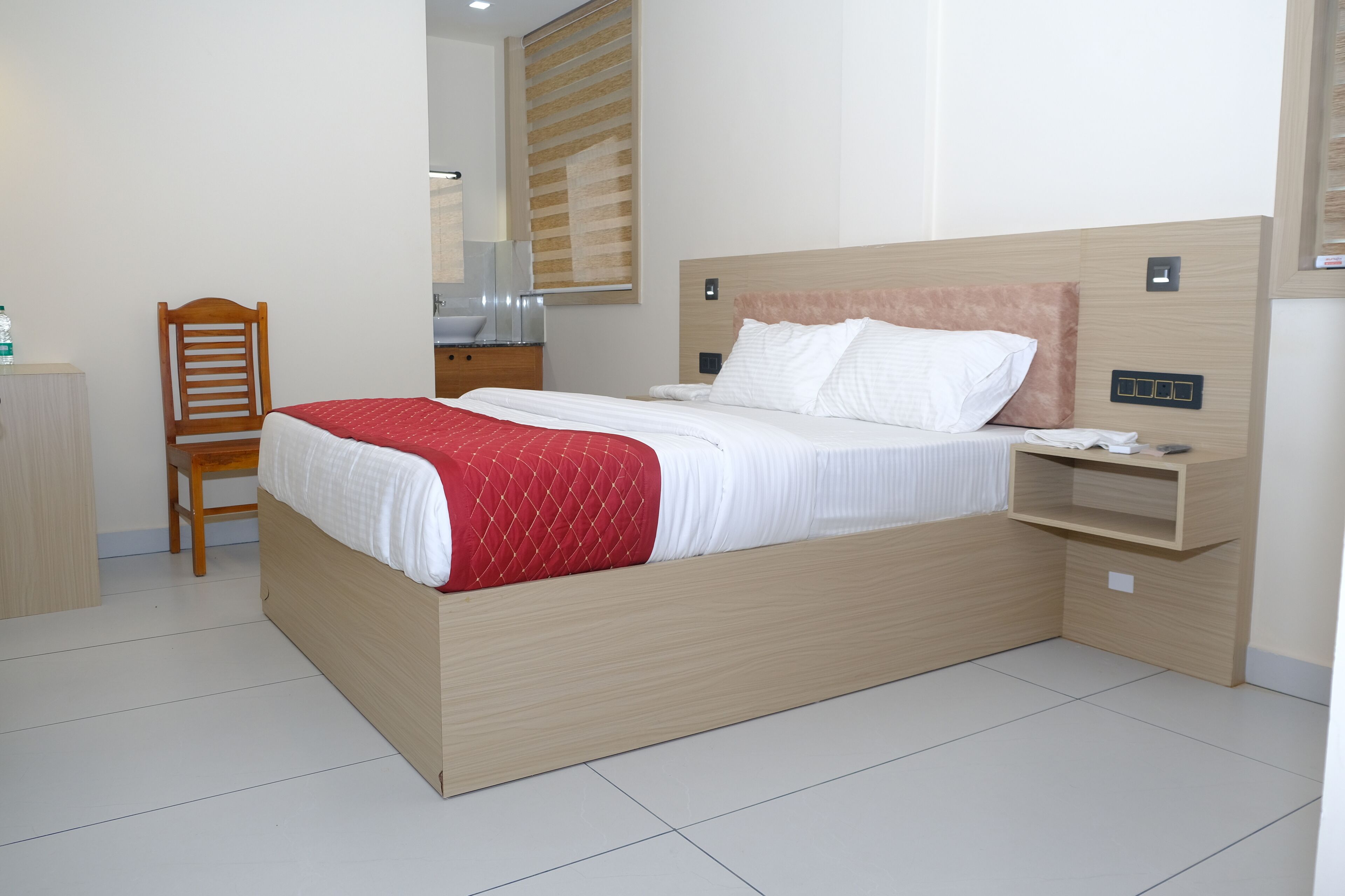 A hotel room with a bed, a wooden chair, a nightstand, and a wall-mounted power outlet.