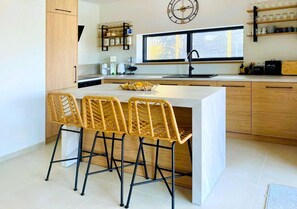 A modern kitchen with a central island, bar stools, wooden cabinets, a clock, and a window providing natural light. - Vila Esnella – Private Pool & Panoramic Sea View (Primošten)