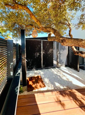 Área externa coberta com deck de madeira, cerca de metal e árvore que oferece sombra.