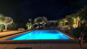 A rectangular swimming pool with a clear blue water surface, surrounded by a paved deck and illuminated by ambient lighting.