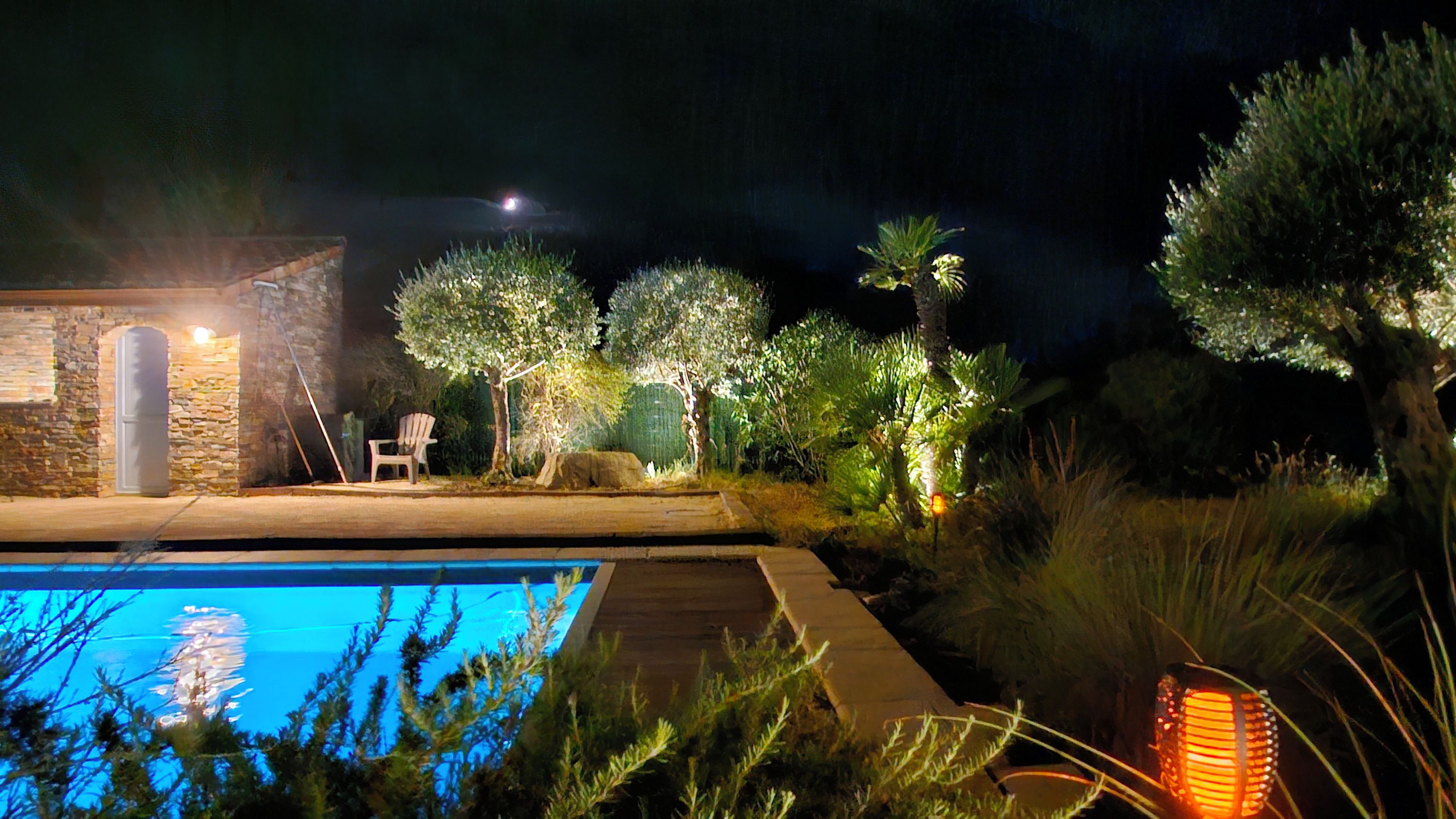 Un espace extérieur aménagé pour la nuit, avec une piscine, des murs en pierre et différents arbres.