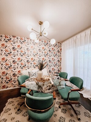 Uma área de refeições com papel de parede floral, mesa de vidro redonda e cadeiras estofadas em verde.