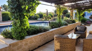 Zona junto a la piscina con muro de piedra, mobiliario de mimbre y abundante vegetación.