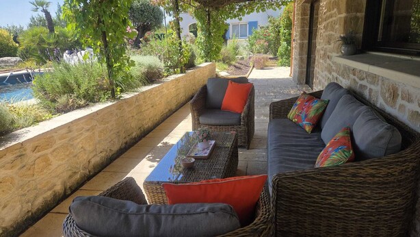 An outdoor seating area with wicker furniture and cushions, a table with a vase, and a view of a pool and greenery.