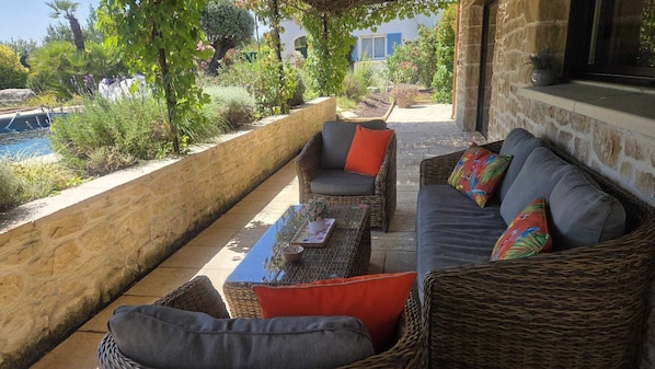An outdoor seating area with wicker furniture and cushions, a table with a vase, and a view of a pool and greenery.