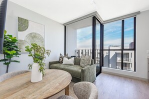 A modern living room with a round wooden table, a green sofa, and a large window offering a city view. - Contemporary Comfort with Natural Elegance (Johannesburg)