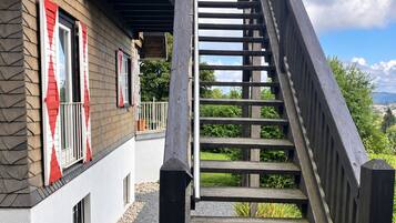 Een houten trap die leidt naar een balkon met uitzicht op groen en een gebouw met een stenen gevel.