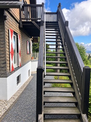 Un escalier en bois mène à un balcon d’où l’on peut admirer la verdure et un bâtiment à la façade en pierre.