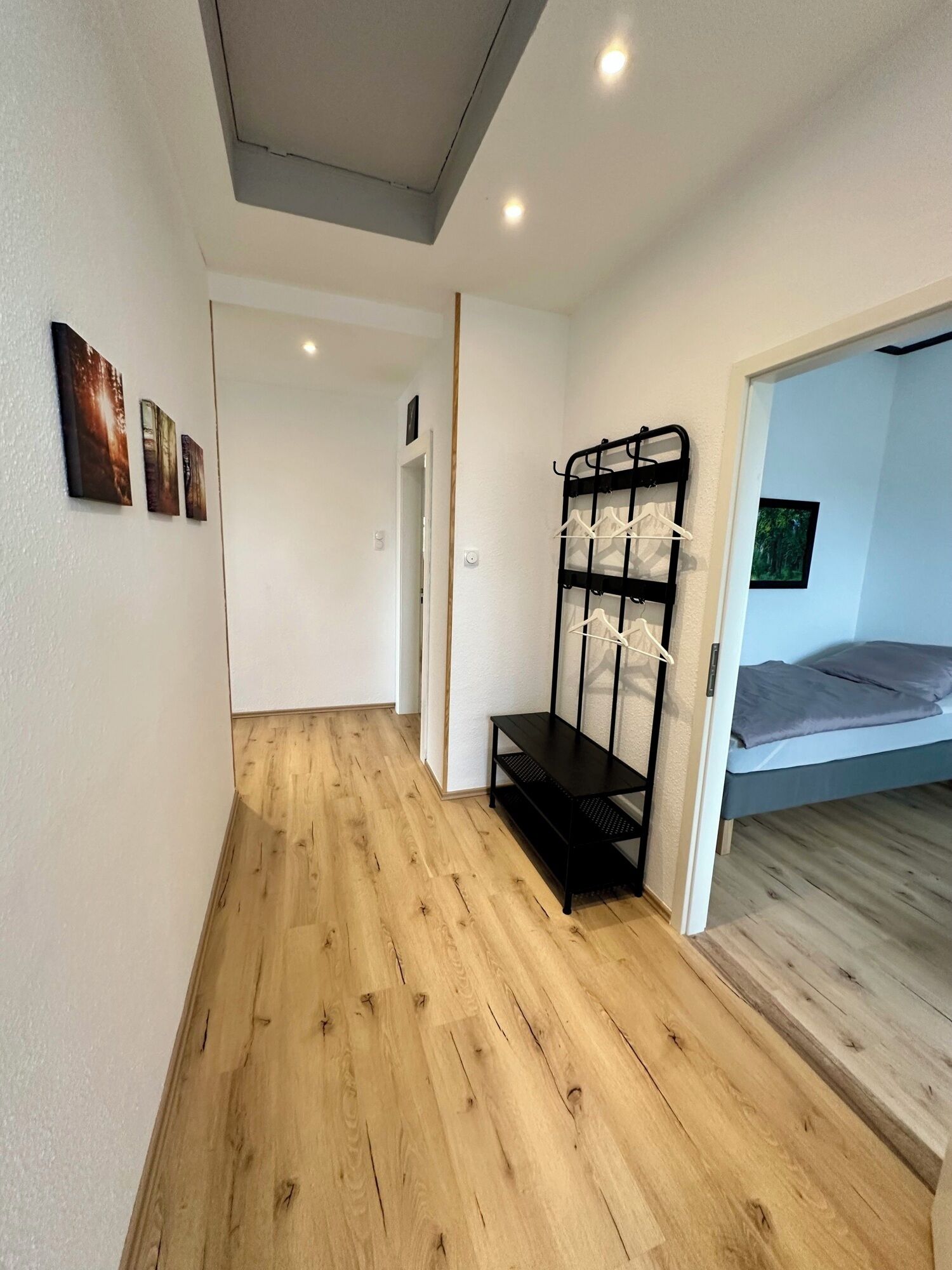 A room with a wooden floor, a bed with grey bedding, a black metal rack with white hangers, and a door leading to another room.
