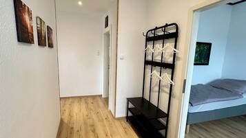 A room with a wooden floor, a bed with grey bedding, a black metal rack with white hangers, and a door leading to another room.