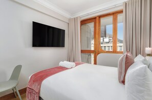 Een hotelkamer met een bed, een aan de muur bevestigde televisie, een raam met gordijnen en uitzicht op gebouwen.