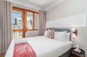A hotel room with a large bed, a nightstand, a wall-mounted light, and a view of buildings through the window. - Charlie and the Chairman | City Views and Style (Cape Town)