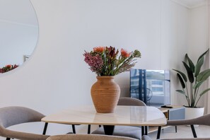 A modern interior with a round table, two chairs, a large vase with flowers, a potted plant, and a television. - Luxe 2xBR Apartment | Ocean Views | Aircon (Umhlanga)