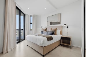 A modern bedroom with a large bed, a nightstand, a lamp, and a view through a large window. - Luxe 2xBR Apartment | Ocean Views | Aircon (Umhlanga)