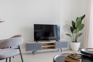 Een moderne woonkamer met een flatscreen televisie op een houten standaard, een grijze bank, een grote potplant en een kleine tafel met boeken en een decoratieve kikker.