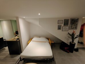 Et hotelværelse med seng, skrivebord, stol, en potteplante og billeder på væggene.