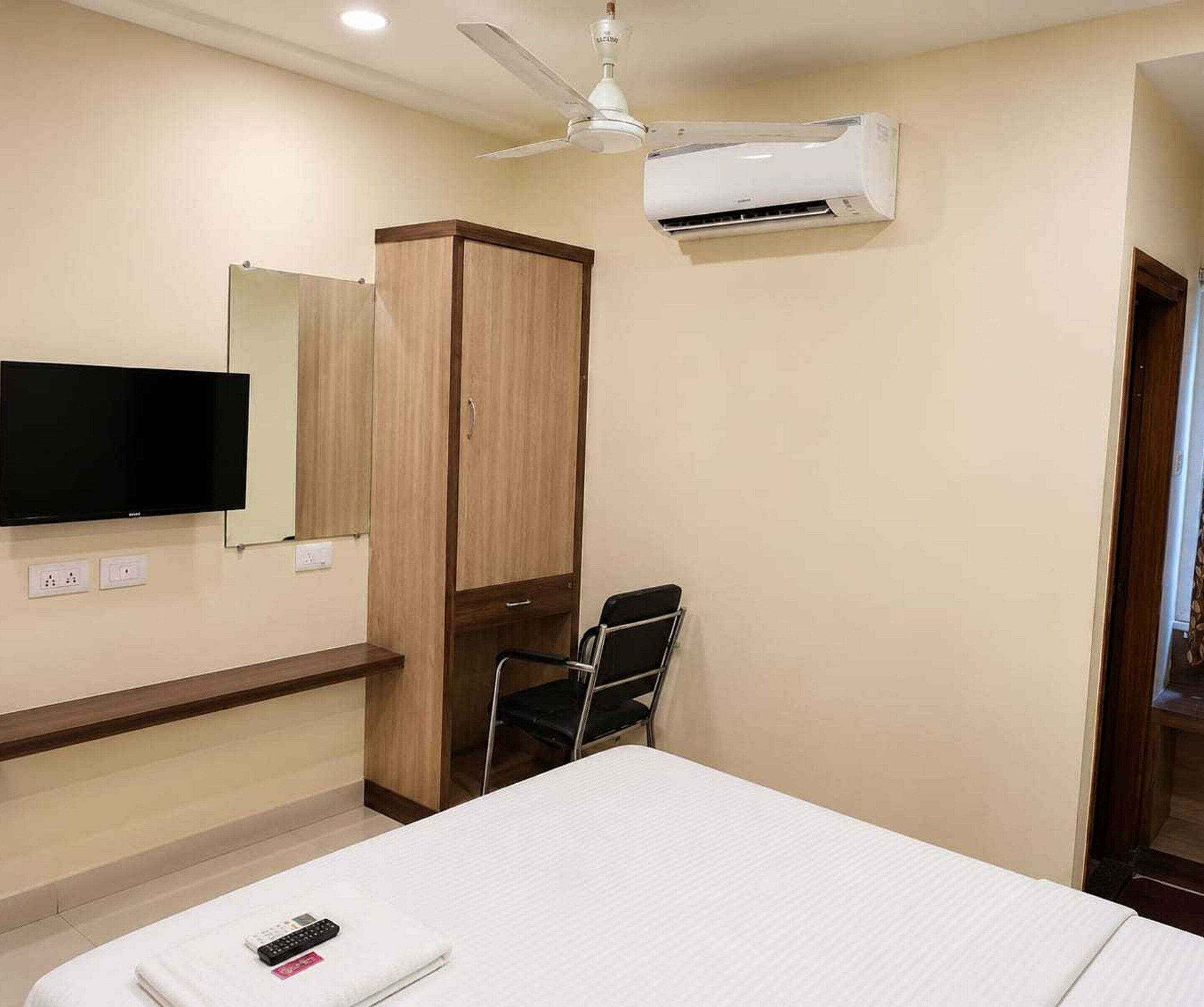 A hotel room with a bed, a TV mounted on the wall, a wardrobe, a chair, and an air conditioning unit.