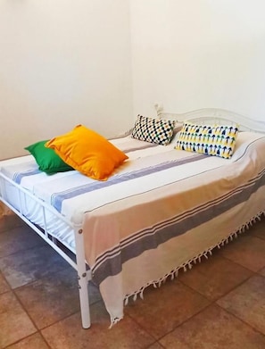Un letto con una struttura in metallo bianco, una coperta a righe e diversi cuscini, tra cui uno verde, uno arancione e uno fantasia.