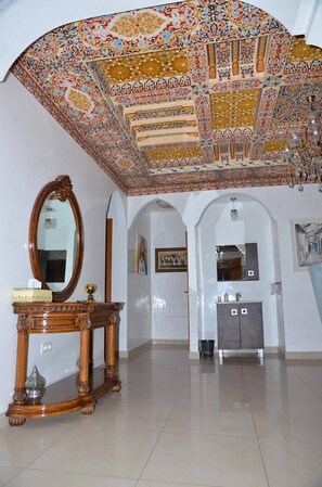 Une pièce dotée d’un grand plafond orné, d’une table en bois avec un miroir, et d’une salle de bain moderne avec un lavabo et un meuble.