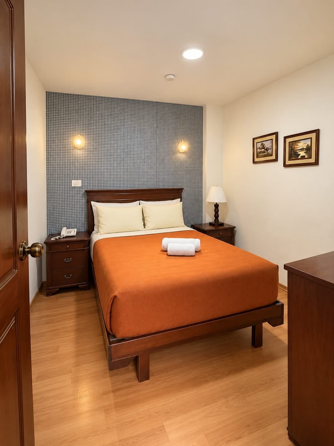 A hotel room with a wooden bed, a bedside table with a lamp, a telephone, and a wall with two framed pictures.