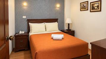 Habitación de hotel con una cama de madera, una mesita de noche con lámpara, un teléfono y una pared con dos cuadros enmarcados.
