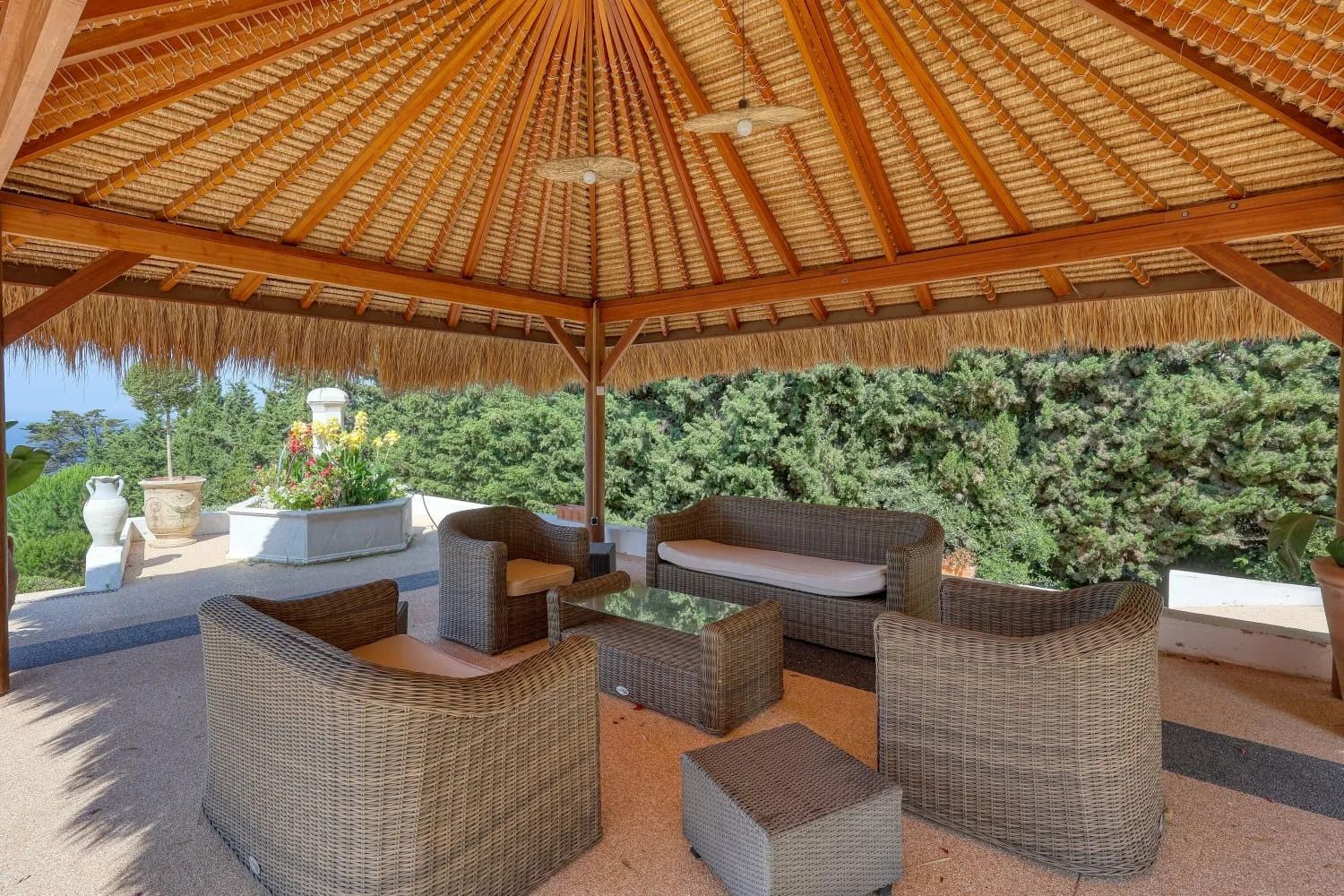 Um pátio externo com móveis de vime, um telhado de palha e vista para um conjunto de sebe verde.