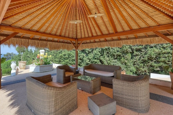 An outdoor patio with wicker furniture, a thatched roof, and a view of a green hedge.