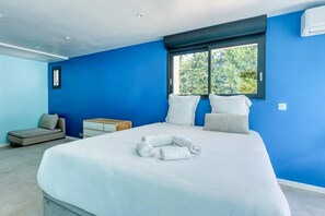 A modern bedroom with a large bed, a window with a view of greenery, a small sofa, and a nightstand.