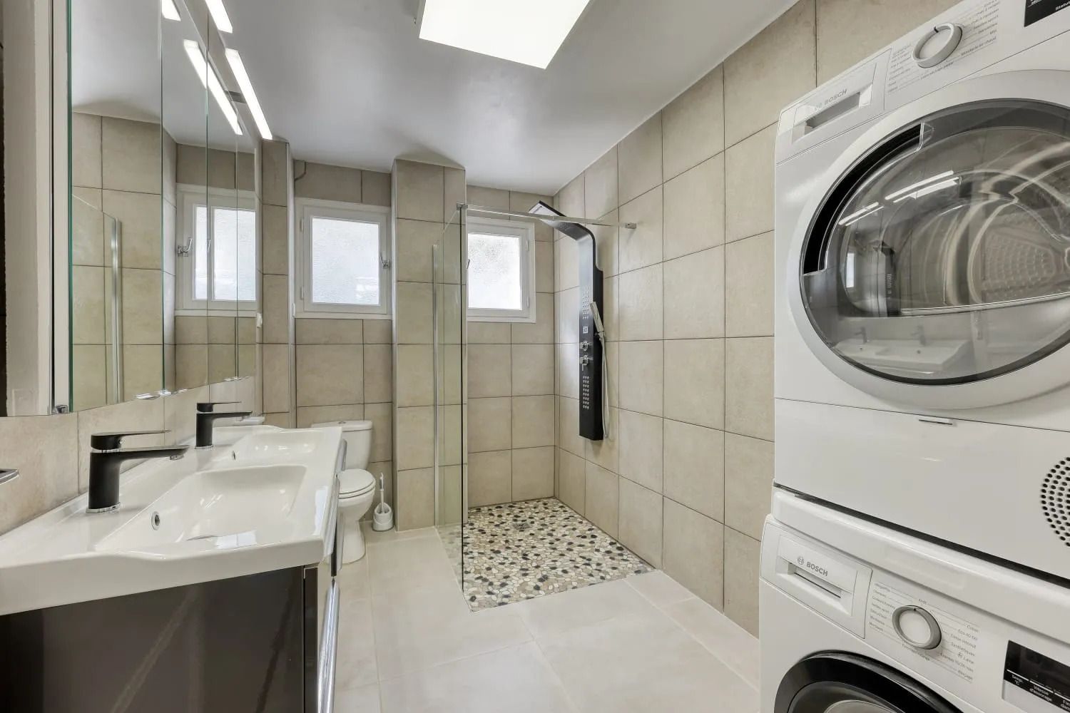 A modern bathroom with a built-in washing machine, a glass shower enclosure, and a large mirror.
