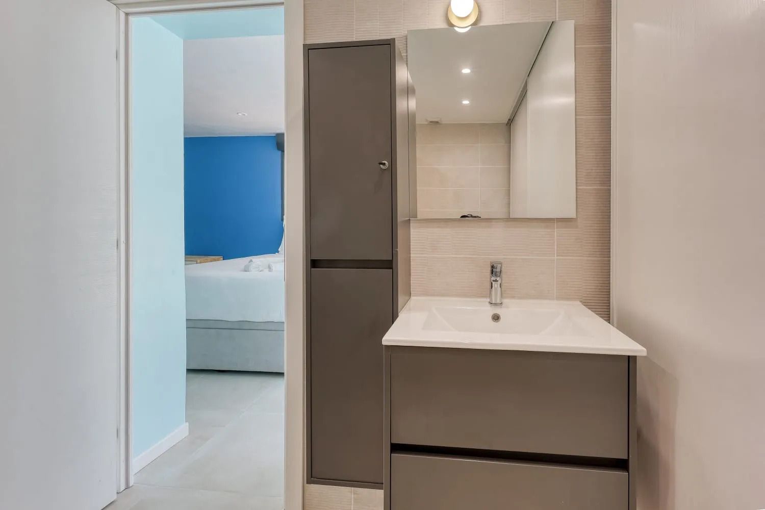 Un baño moderno con un lavamanos blanco, un espejo y una puerta que da a un dormitorio con pared azul.