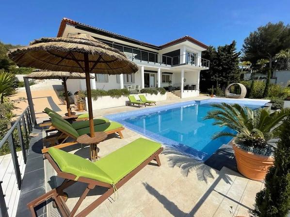 A poolside area with lounge chairs, a thatched umbrella, and a modern house in the background.
