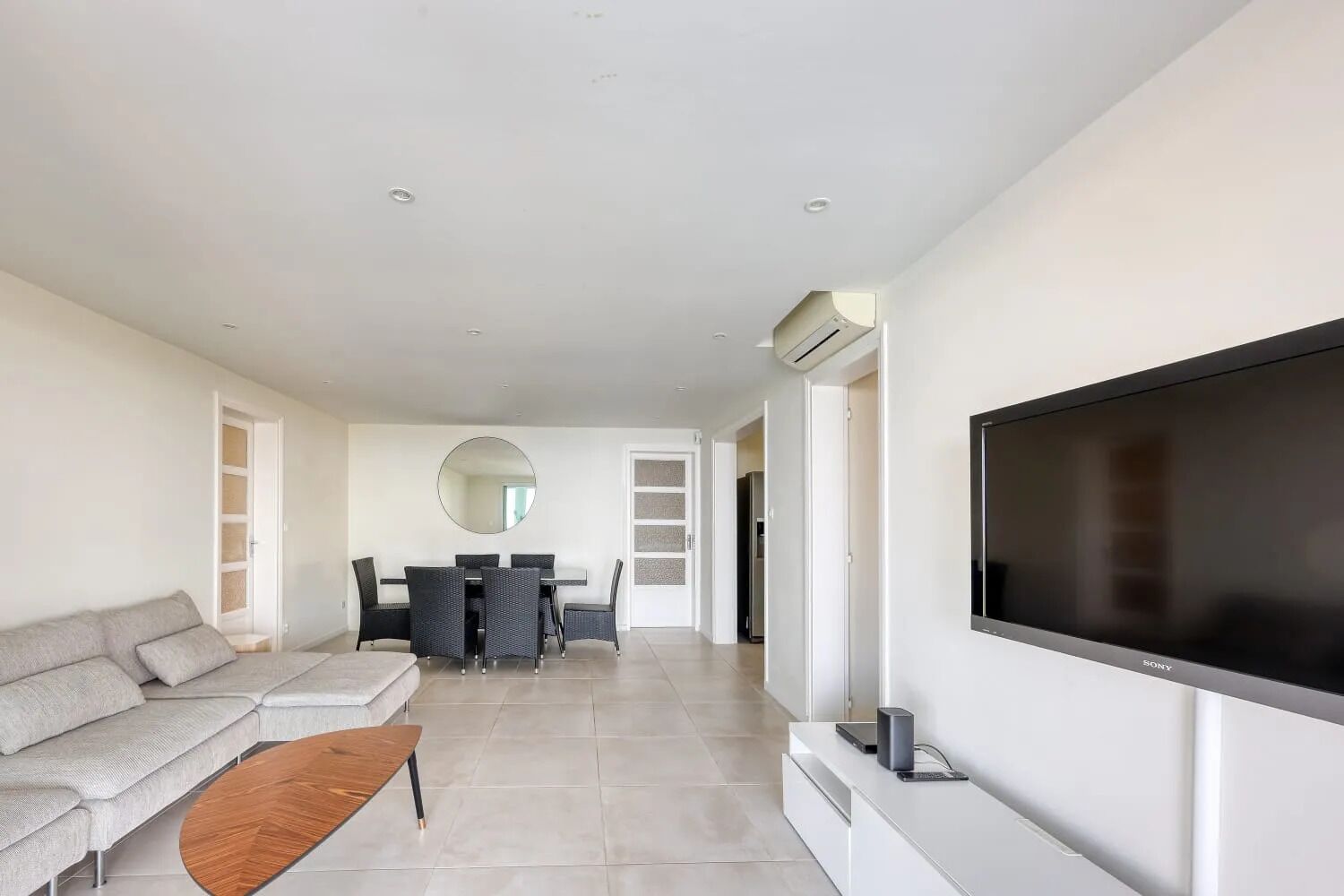 Uma sala de estar moderna com sofá, mesa de centro, TV, área de jantar com cadeiras e um espelho grande.