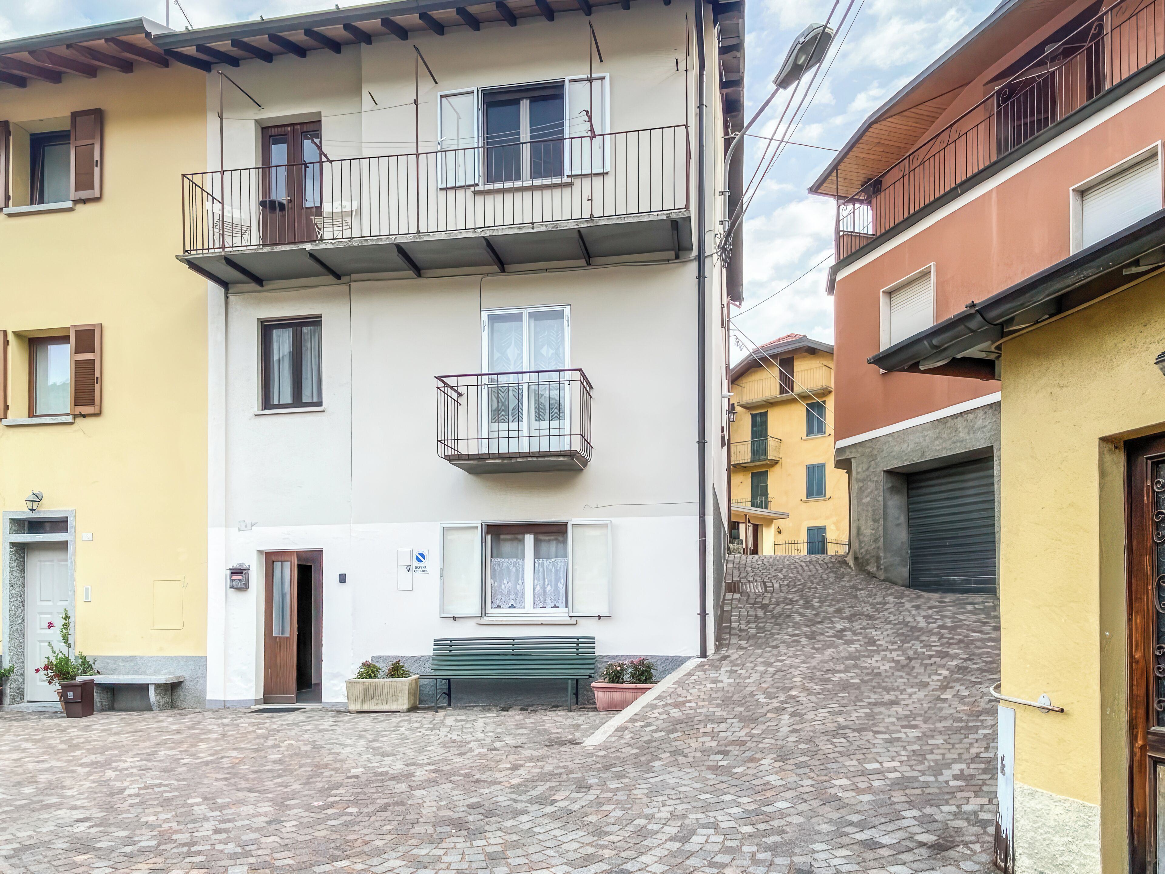 Une ruelle étroite pavée, bordée d’immeubles à plusieurs étages dotés de balcons, avec un banc et des plantes en pot.