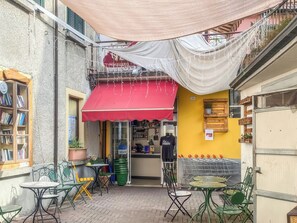 An outdoor café with tables and chairs, a red awning, and a visible storefront with a sign. - Studio Klaus - Schignano (Occagno)