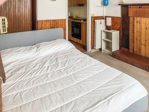 A bedroom with a large bed, wooden paneling, a kitchen in the background, and a small shelf with a lamp. - Studio Klaus - Schignano (Occagno)
